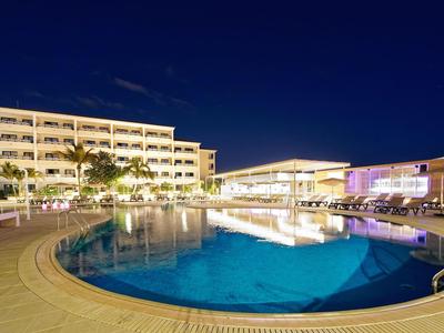 Nachtszene eines beleuchteten Hotels mit großem, rundem Swimmingpool und Palmen.