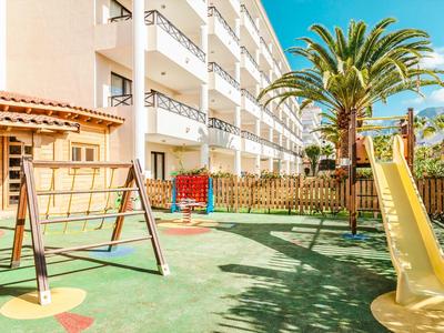 Kinderspielplatz mit Rutsche, Schaukel, Klettergerüst und Palme neben einem weißen Gebäude.