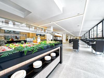 Moderne, helle Kantine mit großem Essensbuffet links und Sitzplätzen rechts, breite Fensterfront.