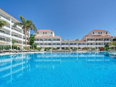 Großer blauer Pool vor modernen weißen Ferienwohnungen unter klarem blauem Himmel.