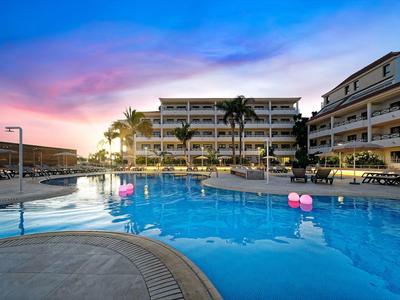 Schwimmbad mit rosa Luftballons vor mehrstöckigem Hotelgebäude bei Sonnenuntergang.