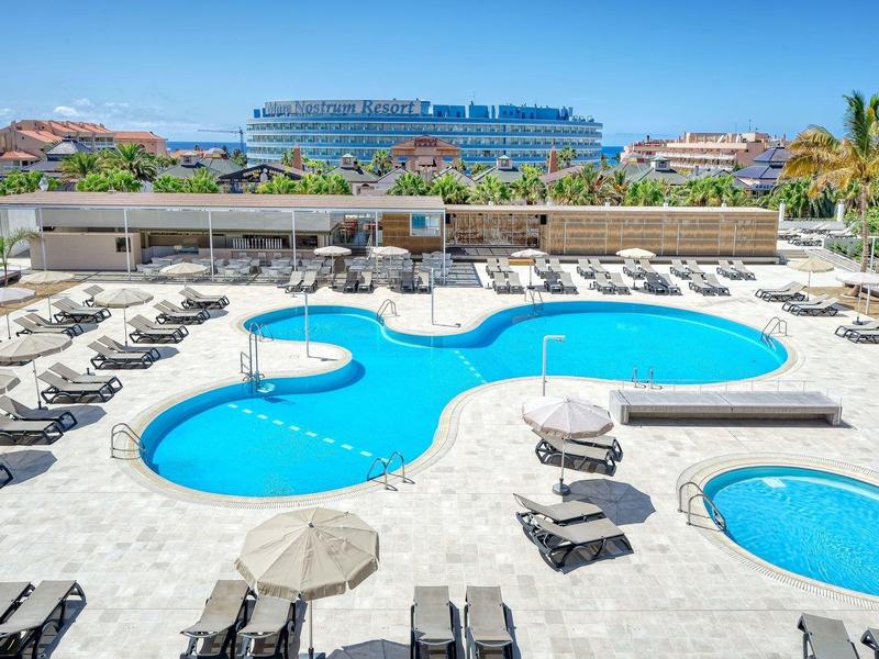 Großer blauer Pool mit Liegestühlen, Sonnenschirmen, Palmen und Gebäuden im Hintergrund.