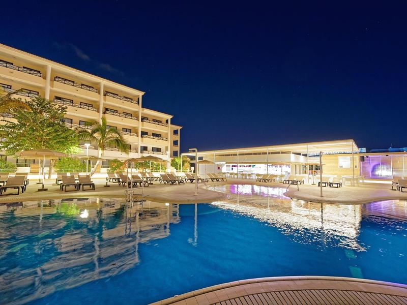 Hotelgebäude und beleuchteter Pool bei Nacht mit klarem Himmel und Spiegelung im Wasser.