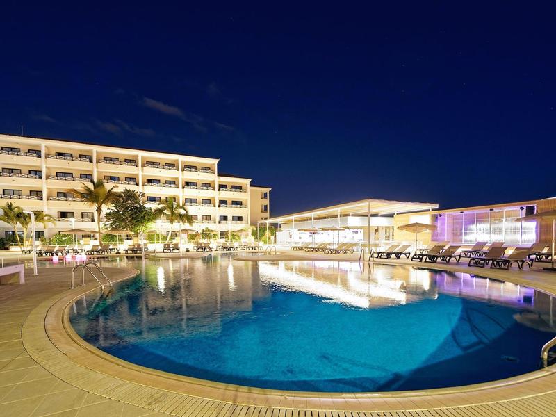 Nachtszene eines beleuchteten Hotels mit großem, rundem Swimmingpool und Palmen.