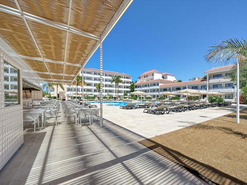 Moderner Hotelpool mit Liegestühlen, Sonnenschirmen, Palmen und hellen Gebäuden unter blauem Himmel.