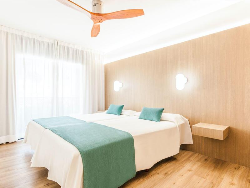 Helles Schlafzimmer mit Doppelbett, hölzerner Wand, blauen Kissen und Decken sowie Deckenventilator.