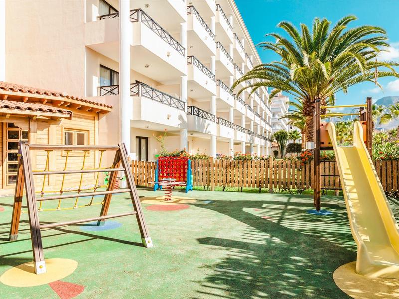Kinderspielplatz mit Rutsche, Schaukel, Klettergerüst und Palme neben einem weißen Gebäude.