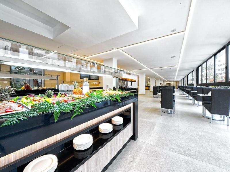 Moderne, helle Kantine mit großem Essensbuffet links und Sitzplätzen rechts, breite Fensterfront.