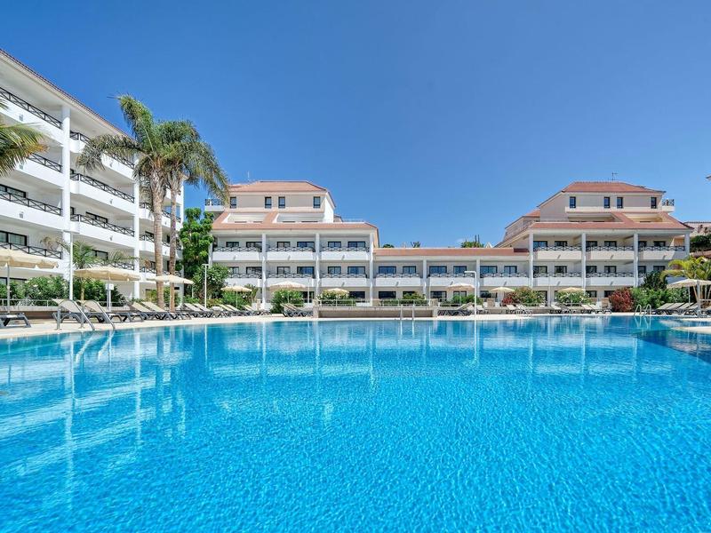 Großer blauer Pool vor modernen weißen Ferienwohnungen unter klarem blauem Himmel.