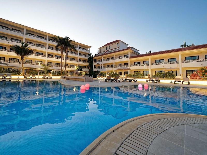 Großer Außenpool mit klar blauem Wasser vor mehrstöckigem Hotelgebäude und Palmen.