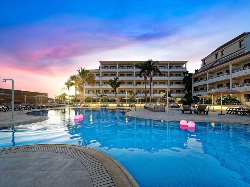 Schwimmbad mit rosa Luftballons vor mehrstöckigem Hotelgebäude bei Sonnenuntergang.