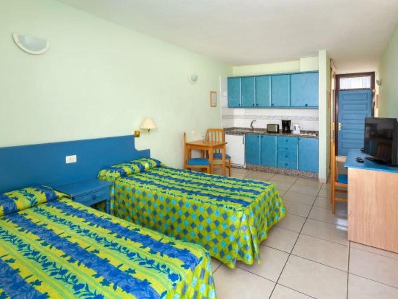 Camera d'hotel con due letti singoli, copriletti blu e verdi, angolo cottura e TV.