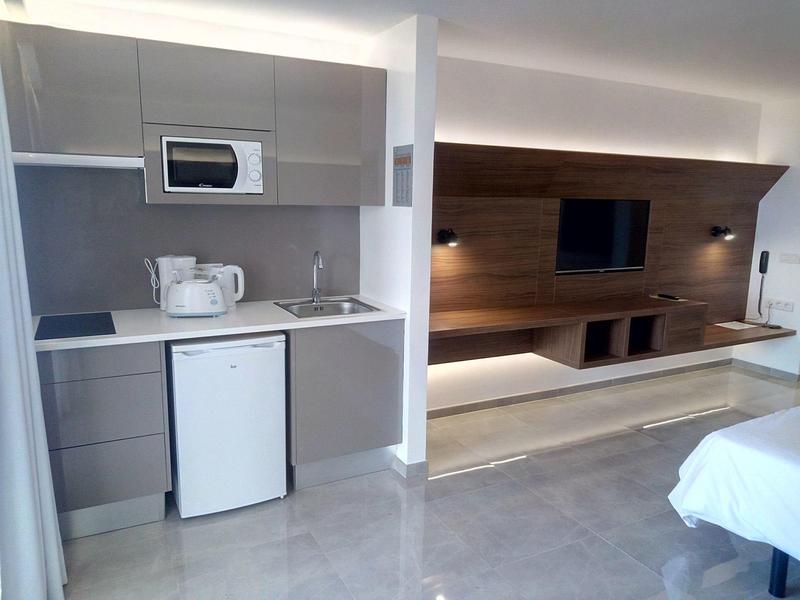 Area camera d'hotel moderna con angolo cottura, microonde, frigorifero e salotto TV con illuminazione indiretta.