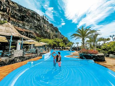Ein Paar geht im Pool vor einer felsigen Berglandschaft mit Palmen und Sonnenschirmen spazieren.