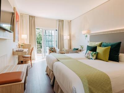 Jasny, nowoczesny pokój hotelowy z dwoma łóżkami, telewizorem i balkonem z widokiem na zieleń.