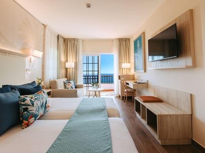 Helles Hotelzimmer mit Doppelbett, blau-grüner Bettwäsche, Fernseher, Schreibtisch und Meerblick durch Fenster.