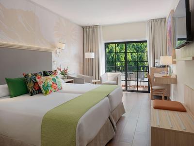 Jasny pokój hotelowy z dwoma łóżkami, zielonymi dekoracjami i balkonem z widokiem na zieleń.