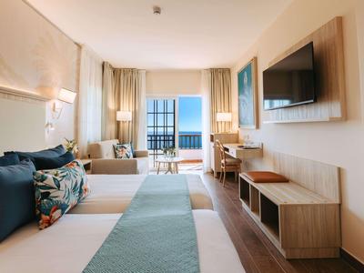 Nowoczesny pokój hotelowy z podwójnym łóżkiem, częścią wypoczynkową, biurkiem i widokiem na morze przez duże okna.