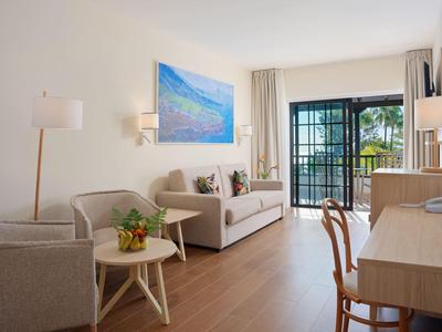 Jasny salon z beżową sofą, fotelami, biurkiem i wyjściem na taras z widokiem na zewnątrz.