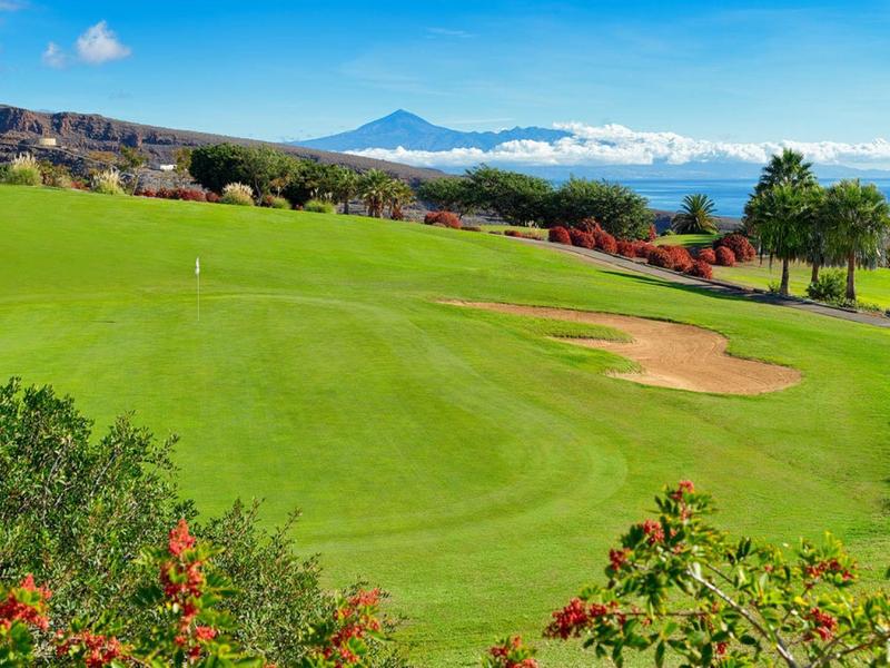 Zielony golf z bunkrem, krzewami i górą w tle pod niebieskim niebem