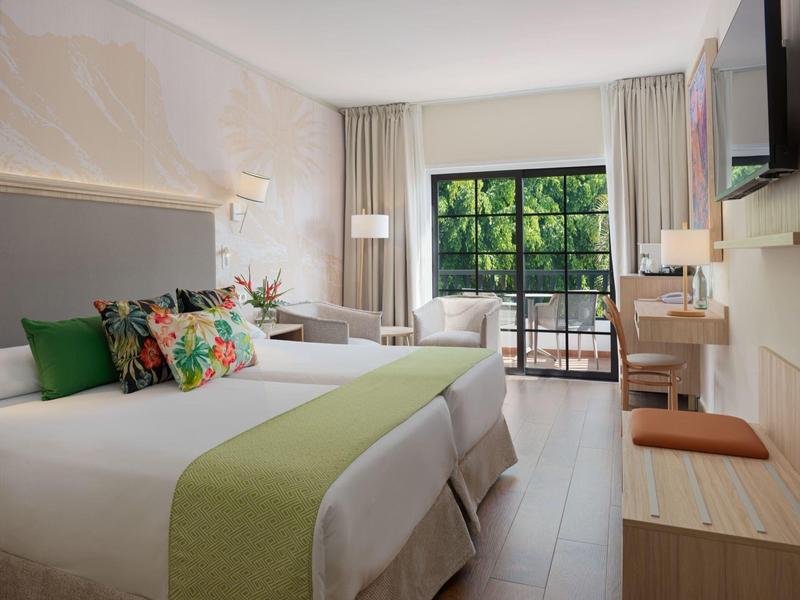 Jasny pokój hotelowy z dwoma łóżkami, zielonymi dekoracjami i balkonem z widokiem na zieleń.