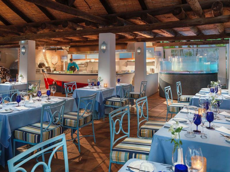 Jasna część restauracyjna hotelu z niebieskimi obrusami i drewnianymi krzesłami pod drewnianym sufitem.