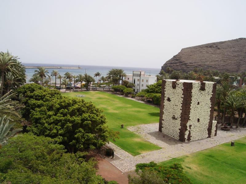 Grüner Park mit einem alten, rechteckigen Steinturm, Palmen und Meer im Hintergrund.