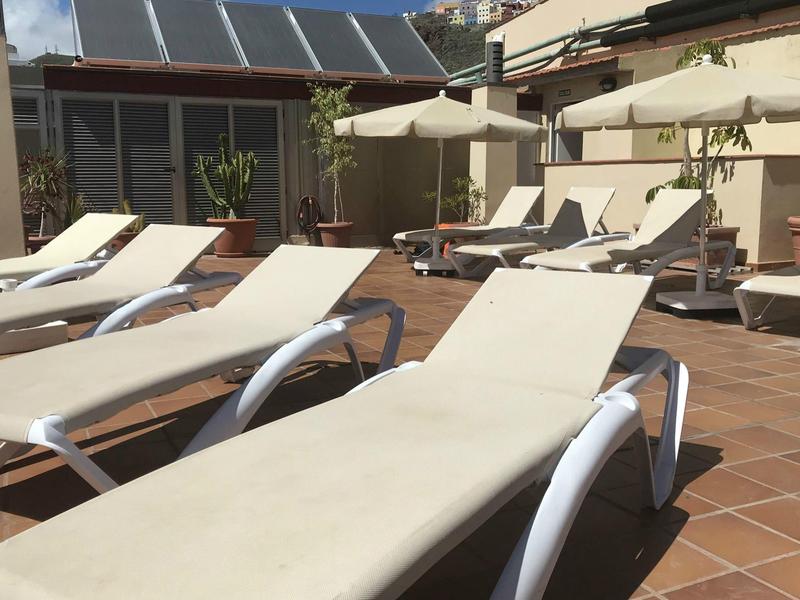 Sonnenliegen mit weißen Polstern und Regenschirmen auf brauner Terrassenfläche unter blauem Himmel.