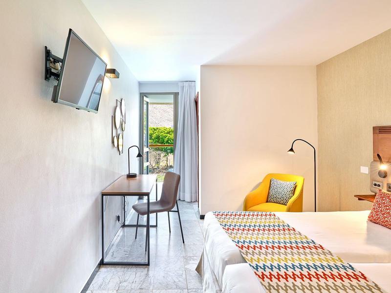 Helles Schlafzimmer mit weißer Wand, Doppelbett, Holzschreibtisch, Stuhl und Balkonblick.