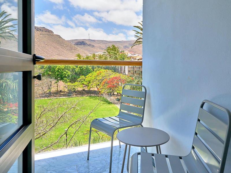Kleiner Balkon mit zwei grauen Stühlen und Tisch, Blick auf grüne Pflanzen und Berge im Hintergrund.