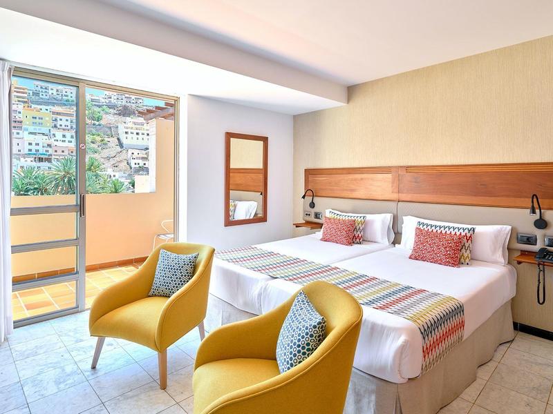 Hotelzimmer mit zwei Einzelbetten, gelben Sesseln und Zugang zu einem Balkon mit Stadtblick.