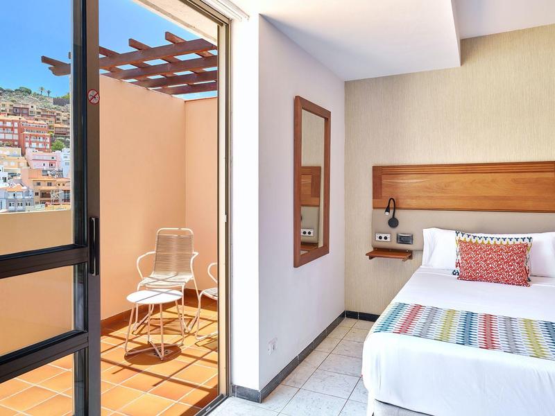 Helles Zimmer mit Einzelbett, buntem Kissen, Holzkopfteil und Balkon mit Stühlen und Tisch.