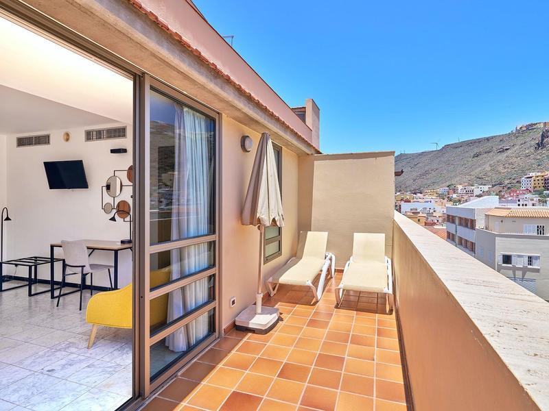 Balkon mit zwei Liegen, Sonnenschirm und Blick auf die Stadt und Berge bei blauem Himmel.