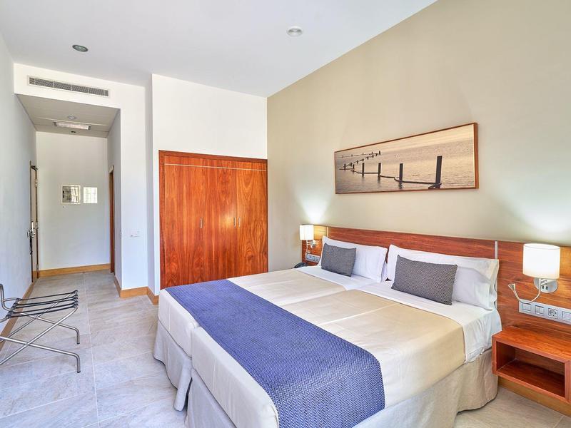 Helles Hotelzimmer mit Doppelbett, blauer Bettdecke, Holzmöbeln und einem Landschaftsbild an der Wand.