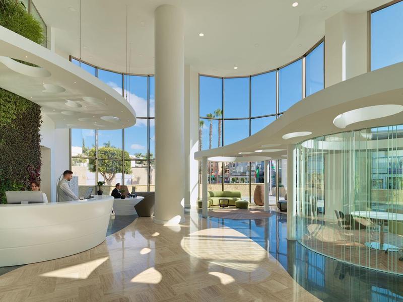 Lichte, moderne hotellobby met hoge ramen, groene muur en strak receptiebalie.