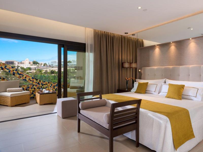 Lichte hotelkamer met groot bed, modern interieur en balkon met zitplaatsen en uitzicht.