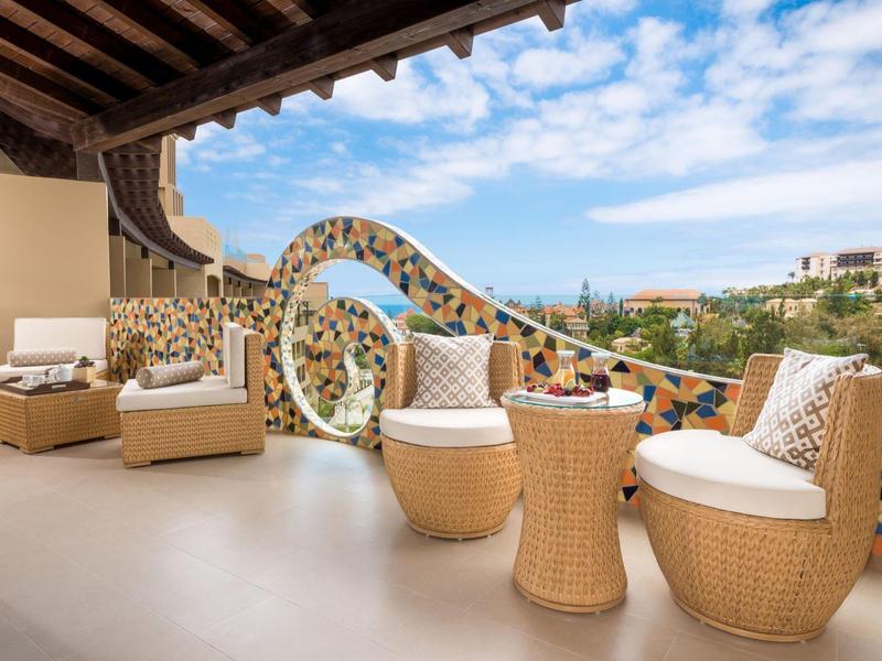 Terras met rotan stoelen en decoratieve balustrade met uitzicht op een stad bij helder weer.