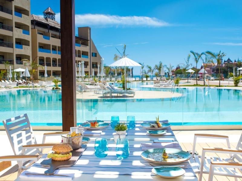 Een gedekte tafel met servies en glazen voor een zwembad en een hotelgebouw.
