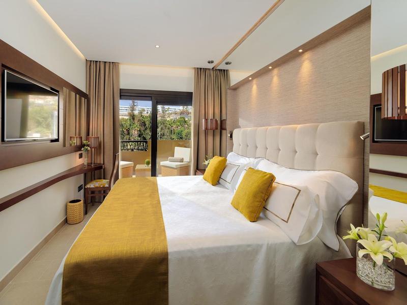 Moderne hotelkamer met tweepersoonsbed, gele accentkussens en balkon met uitzicht op groene omgeving.