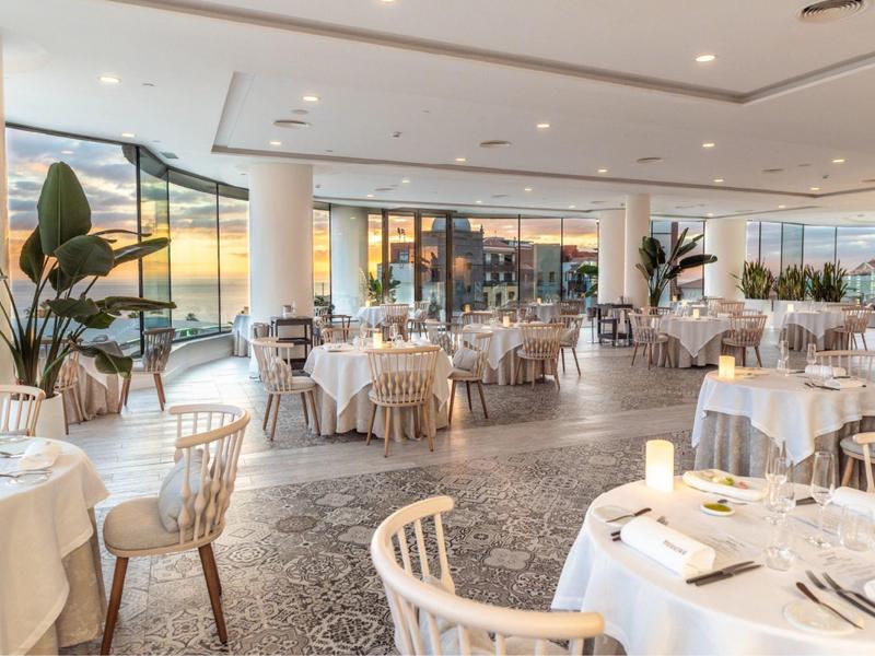 Licht en elegant restaurant met ronde tafels, witte tafelkleden en grote ramen met uitzicht op zee.