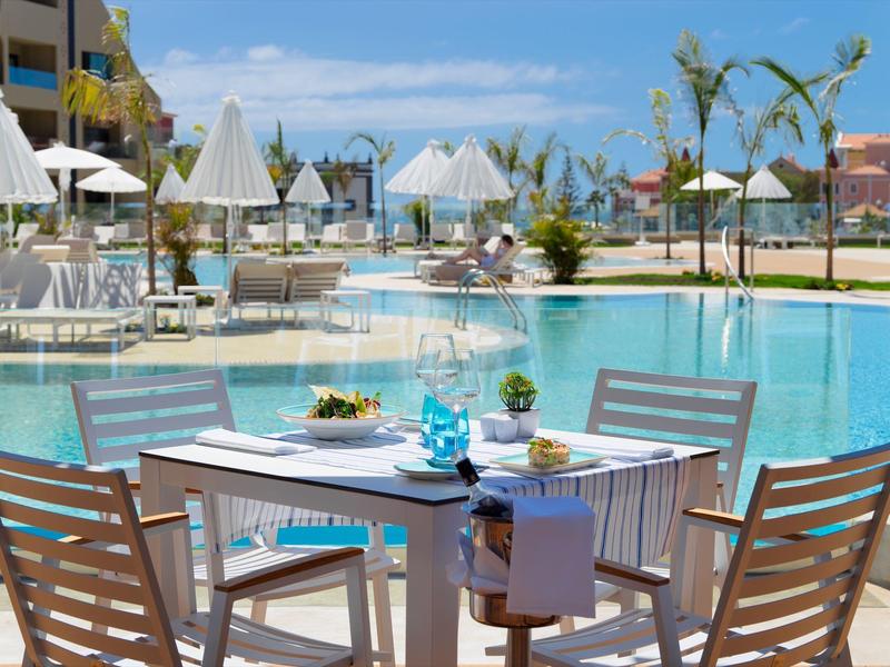 Een gedekte tafel met stoelen naast een hotelzwembad met parasols en palmbomen.