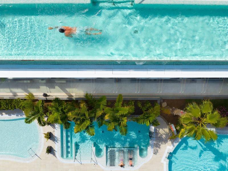 A person swimming in a long pool above a shaded pool area with palm trees.