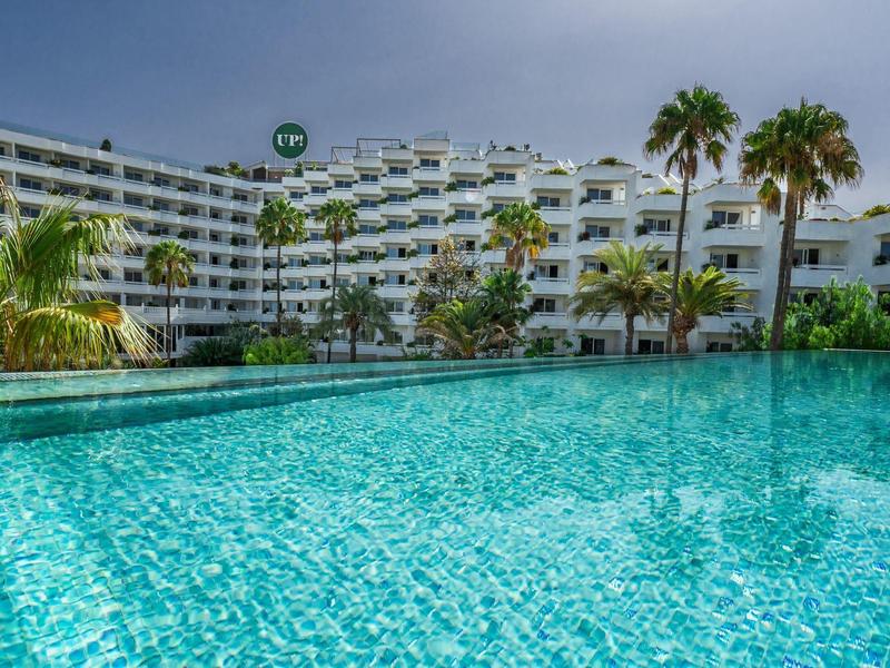 Modern hotel with large pool and palm trees under a cloudy sky.