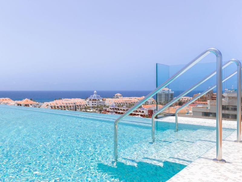 Clear blue pool with metal handrails overlooking ocean and beach umbrellas.