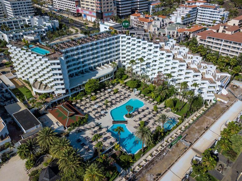 Modern hotel with terraced white balconies surrounding a blue pool and lush palm trees.