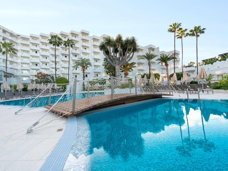 Resort pool area with a wooden bridge, palm trees, and white multi-story buildings.