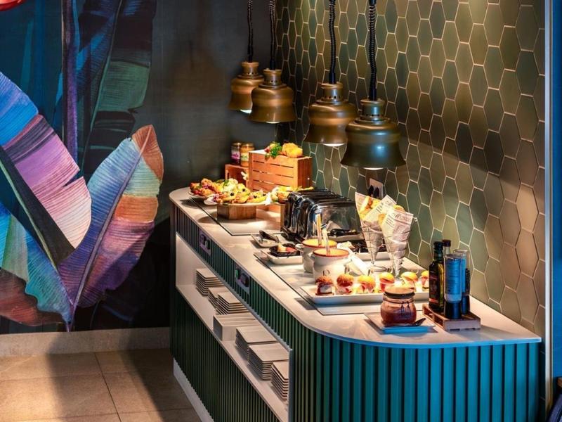 Modern buffet with desserts and drinks in a stylish setting with honeycomb tile wall and colorful artwork.