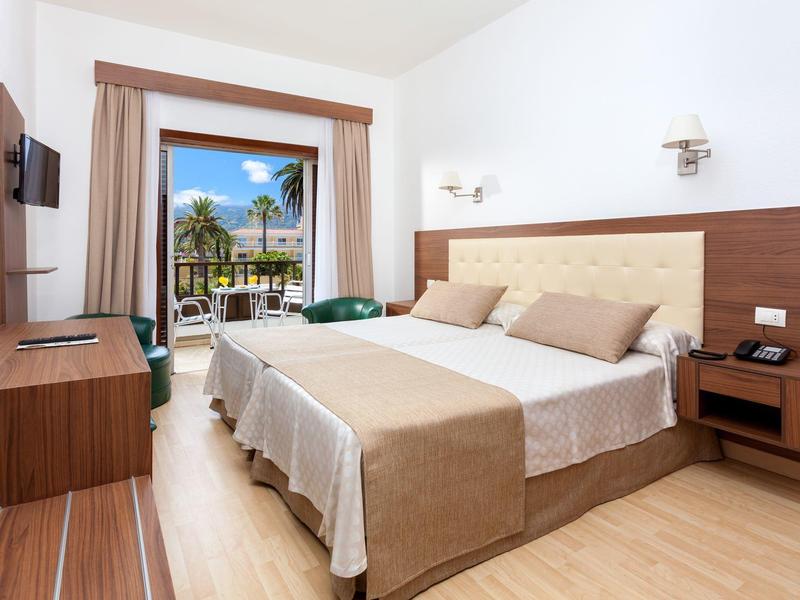 Licht hotelkamer met tweepersoonsbed, houten meubels en uitzicht op terras met palmbomen.