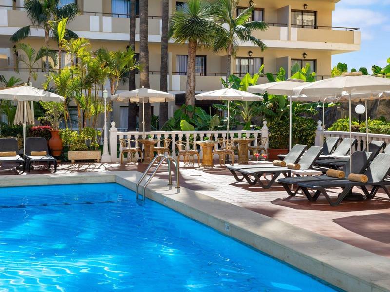 Hotel zwembad met ligstoelen, parasols en palmbomen bij zonnig weer.