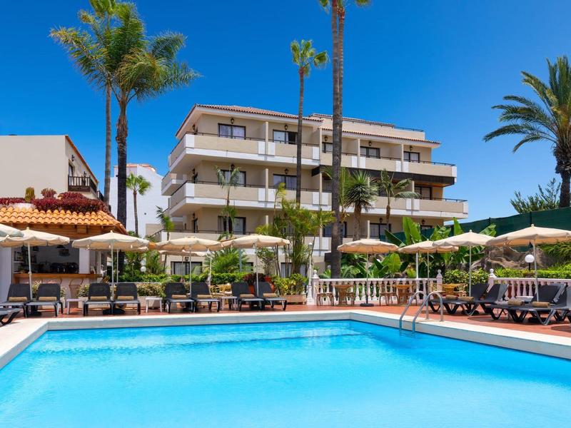 Hotelzwembad met ligstoelen en parasols voor een meerlaagse gebouw en palmbomen.